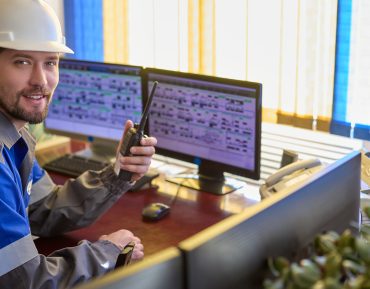 Smiling Caucasian worker in overalls and a helmet uses a walkie-talkie, looks at the camera sitting at desk in the control room. Automated workplace of dispatcher or operator of a modern enterprise